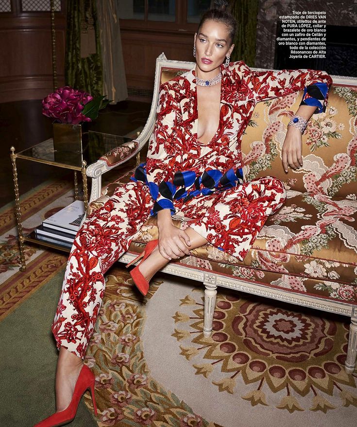the image showcases a fashion model elegantly posed on a vintage armchair in a luxurious setting. she's wearing a striking red and white floral jumpsuit, paired with red highheeled shoes. the model is adorned with exquisite cartier jewelry, including a necklace, bracelet, and earrings. the background features an ornate armchair, a richly patterned carpet, and classical interior design elements, all contributing to a highfashion, editorial feel. the overall atmosphere is one of luxury and sophistication.