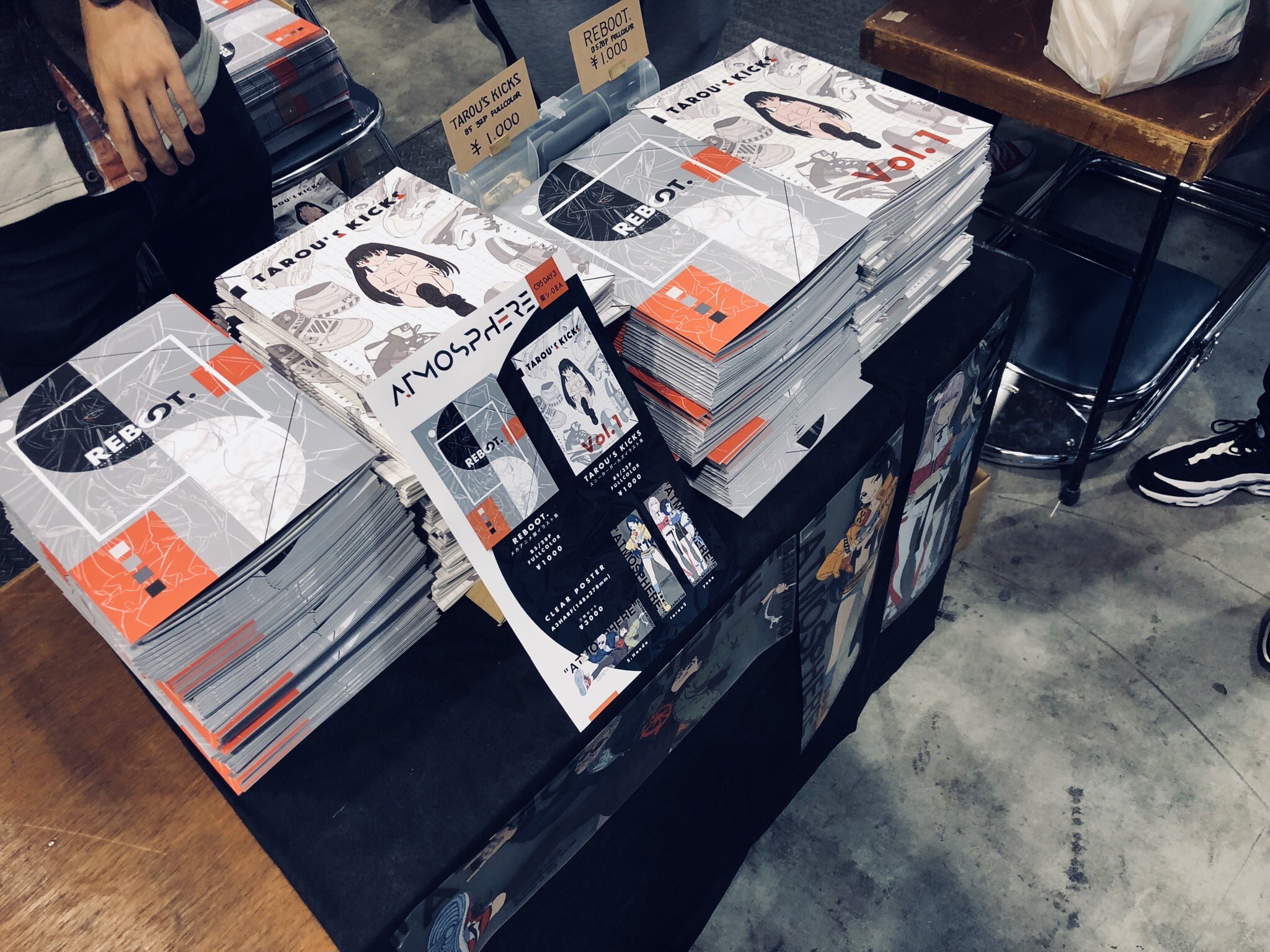 a table filled with stacks of selfpublished manga and art books at a comic market. the table is covered with a black cloth, and there are some promotional flyers and bookmarks on display as well. a person's hand is visible on the left side of the image.