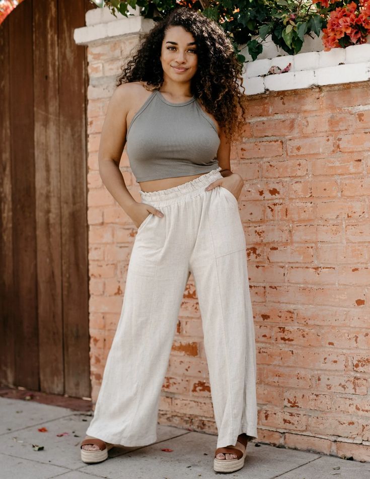 a young woman with curly hair is posing outdoors in front of a brick wall. she is wearing a green halter neck crop top and beige wideleg pants. she is also wearing sandals and has her hands in her pockets. the photo is taken from eye level and the lighting is natural and sunny.