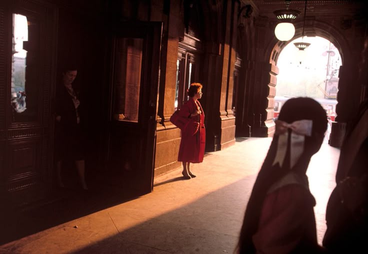 a photograph captures a woman in a red coat standing in a sunlit doorway of an old building. the doorway is part of an archway, leading to a street outside.  another woman, partially visible, is in the background near the doorway, and a girl with long dark hair and a bow is in the foreground, slightly out of focus. the sun casts strong shadows, creating a dramatic contrast of light and dark. the overall aesthetic suggests a vintage or retro style of photography.