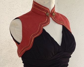 a red cape with gold trim and a high collar is displayed on a mannequin. the cape is secured with an ornamental clasp. the mannequin is wearing a black dress.