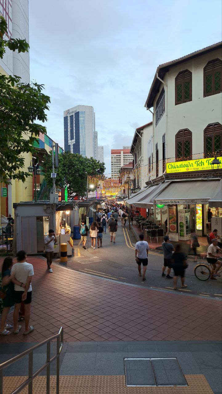 a bustling street scene in singapore, showcasing a mix of old and new architecture. the foreground features a tiled walkway with pedestrians walking by, while the background showcases traditional shophouses with bright yellow signs. the scene is framed by modern skyscrapers and a cityscape in the distance, highlighting the city's unique blend of modernity and heritage.