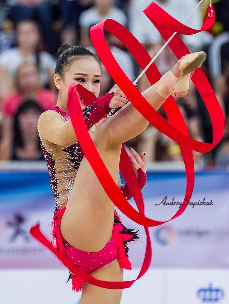 a young female rhythmic gymnast is midperformance, displaying her athleticism and skill. her leg is extended high in the air, as she manipulates a flowing red ribbon with precision. the focus of the image is the gymnast's dynamic pose and the ribbon's movement, highlighting the artistry and grace involved in this sport. the vibrant red ribbon contrasts with the surrounding audience, creating a compelling visual. the gymnast's expression is focused and determined, showcasing her dedication and passion for the sport.