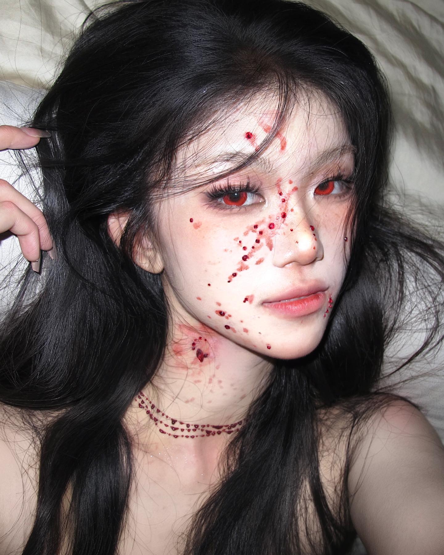 a young woman with long black hair poses for a selfie. she's wearing elaborate halloween makeup, styled to give the appearance of a vampire with fake blood around her mouth and neck. her red eyes, achieved with contact lenses, enhance the vampire effect. she's lying on a white bedspread, and the soft lighting emphasizes the details of her makeup.
