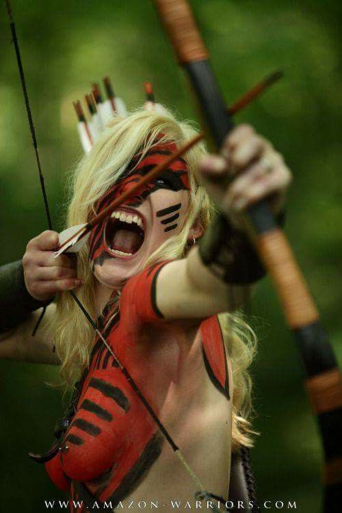 a woman dressed in red body paint as a warrior holds a bow and arrow and looks directly at the camera. the woman is in focus and the background is blurred. the bow and arrow and the woman's face paint are the main subjects of the image.