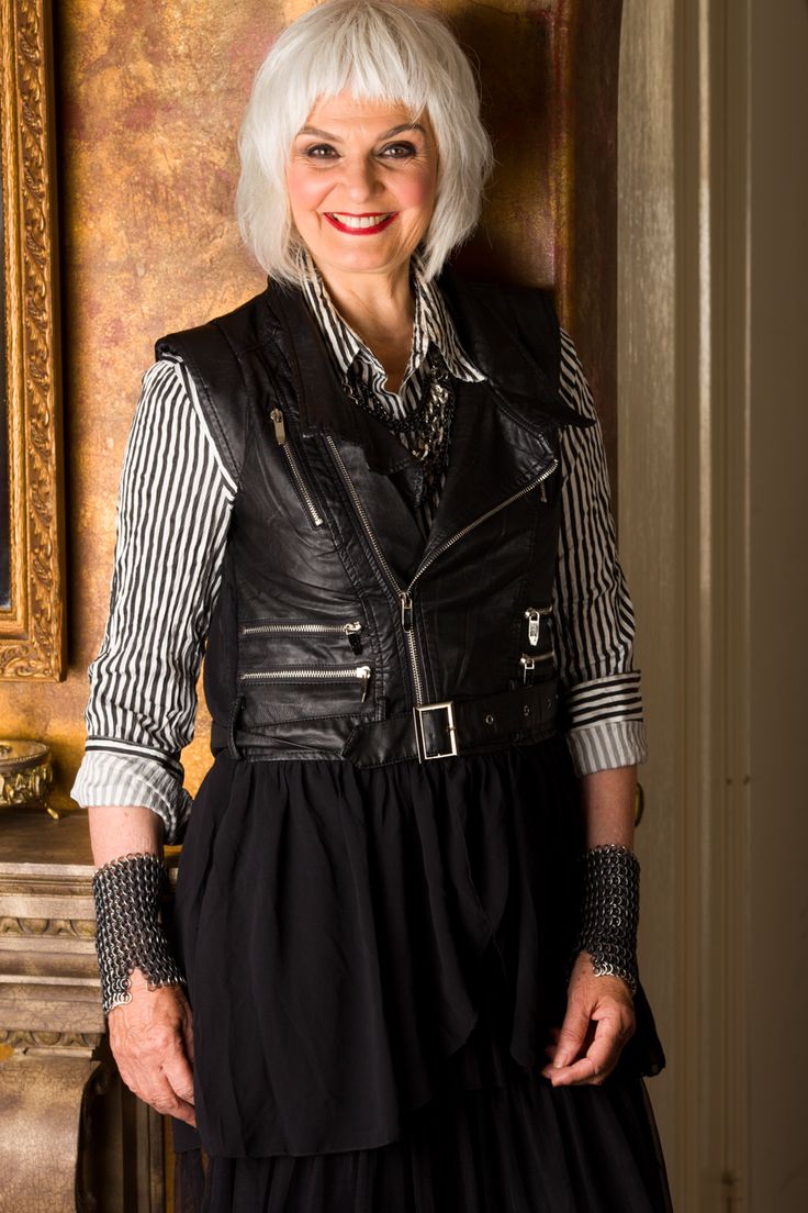 a woman with white hair and a short hairstyle is standing in front of a wall. she is wearing a black leather jacket over a black and white striped shirt, a black skirt, a silver necklace, and chain bracelets. she is smiling and looking at the camera. the background is a wall with a gold frame on the left and a brown door on the right.