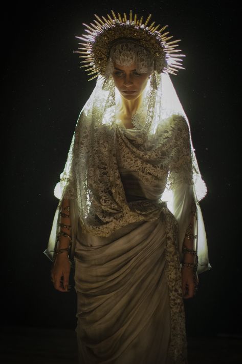 a woman in a long, flowing gown with a lace veil and a headdress resembling a halo or crown of spikes stands against a dark background. the lighting is dramatic, highlighting her face and costume, giving the image an ethereal and somewhat religious feel. she wears jewelry on her arms.