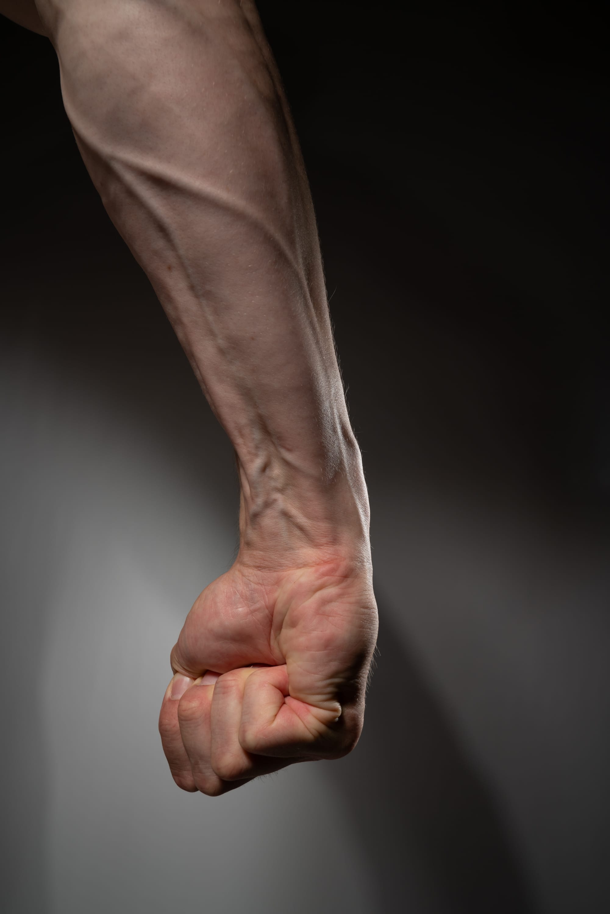 a closeup shot of a muscular man's arm and clenched fist against a gray background. the image emphasizes the strength and tension in the arm, highlighting the prominent veins and tendons.