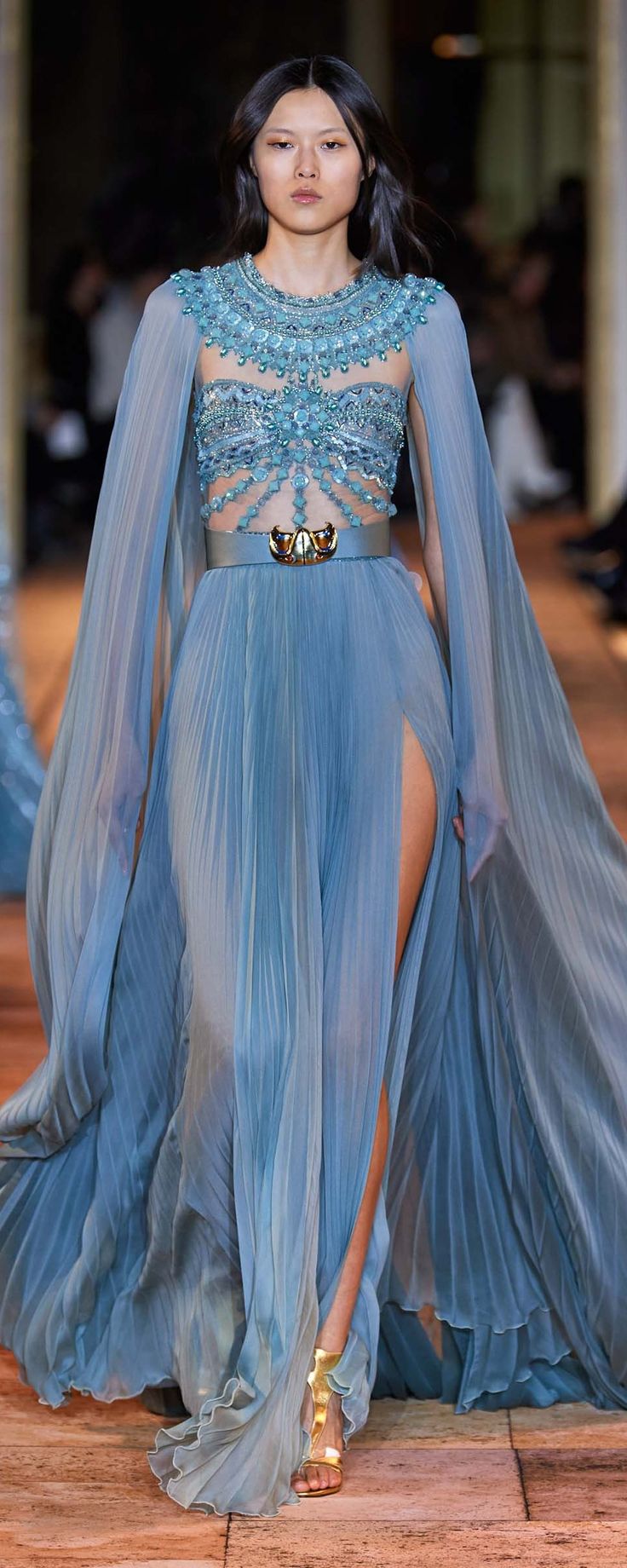 a model walks the runway in a stunning light blue haute couture gown. the dress features a pleated skirt with a high slit, a sheer beaded bodice, and dramatic cape sleeves. a gold belt cinches the waist, and gold sandals complete the look. the model's hair is styled simply, and her makeup is natural. the background is blurred, but shows the typical setting of a fashion show.  the overall impression is one of elegance and sophistication.