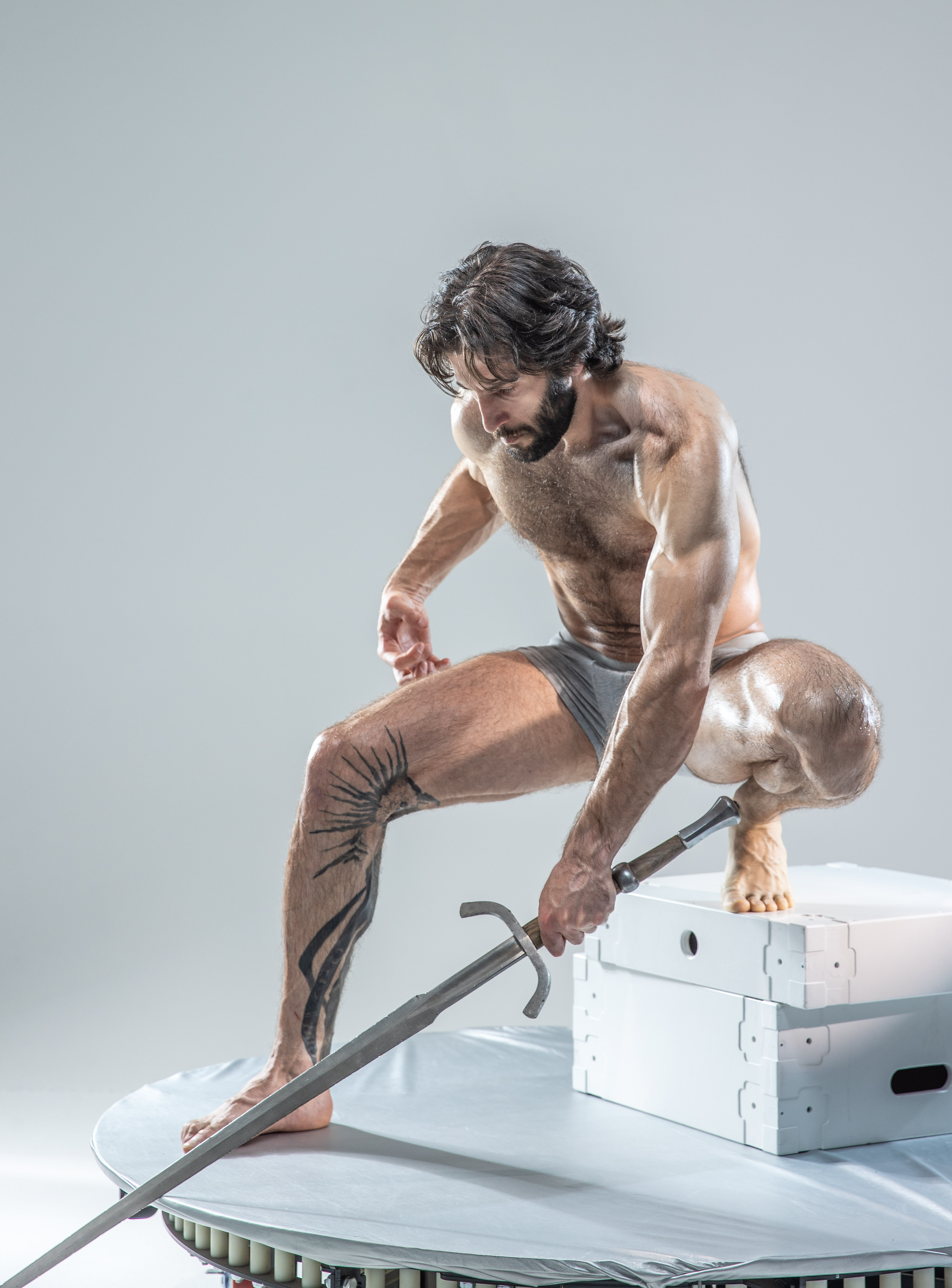 a muscular man with long dark hair and a beard is posed in a dynamic stance on top of and beside white boxes. he is holding a sword in his right hand. he is wearing only athletic shorts and has a tribal tattoo on his left leg. the background is a light gray.