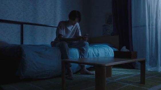 a person is sitting on a bed in a dimly lit room, looking down at their phone. the room is dark and mostly blue, with a single bed and a small table in front of it. the person is wearing jeans and a tshirt and is sitting with their legs crossed. the overall mood of the image is one of sadness and isolation.