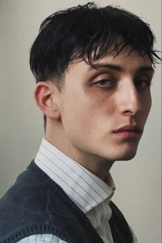 a young man with dark hair and a serious expression is looking directly at the camera. he is wearing a white striped shirt and a dark sweater vest. he is standing indoors against a lightcolored background. the lighting is soft, creating subtle shadows on his face.