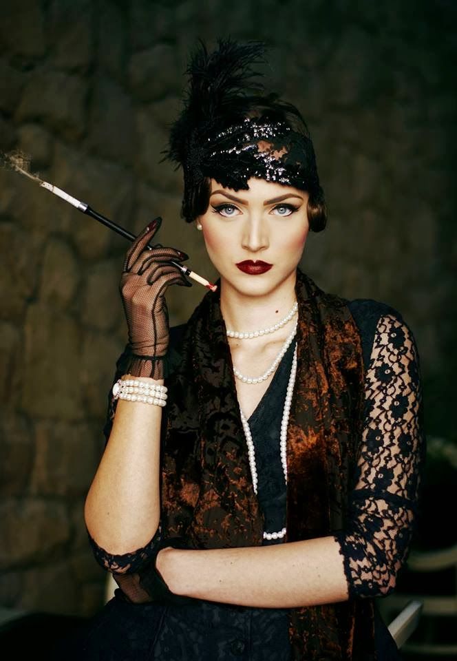 a young woman in a glamorous 1920s flapper style outfit poses with a cigarette and a confident expression. she is wearing a black beaded headband with a feather, black lace gloves, a long pearl necklace, and a dark brown velvet dress with black lace details. her red lipstick and dramatic eye makeup enhance her elegant look.  she appears to be in a studio setting with a dark stone wall backdrop. the image captures a sense of vintage glamour and sophistication.