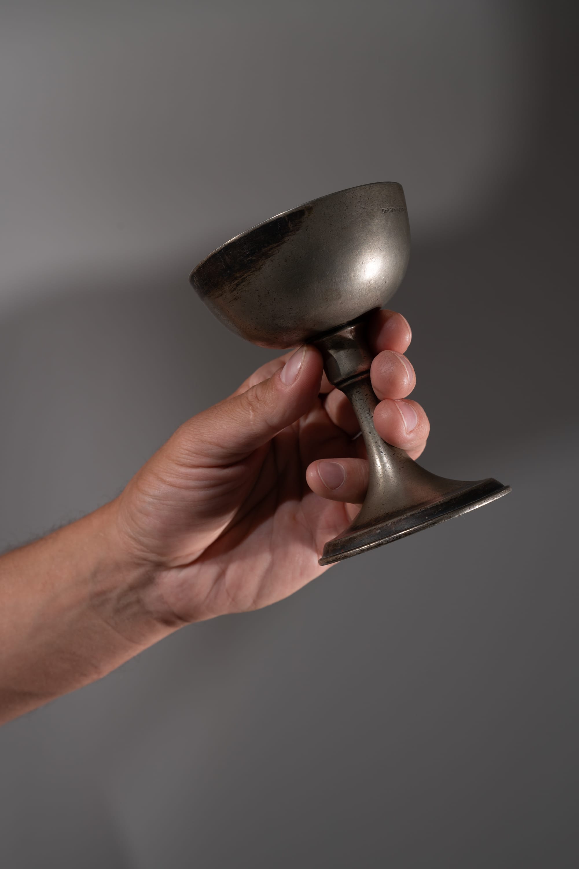 a hand holds an old, tarnished metal cup against a light gray background.