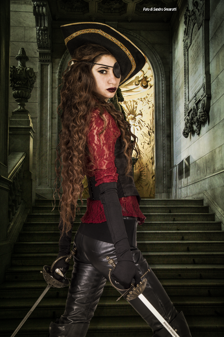 a woman dressed as a pirate stands on a staircase, holding two swords. she is wearing a black pirate hat with gold trim and an eye patch over one eye. her outfit includes a red lace top, a black vest, and tight black leather pants with boots. she gazes back over her shoulder. the background shows a grand staircase and an ornate archway with a gold interior.