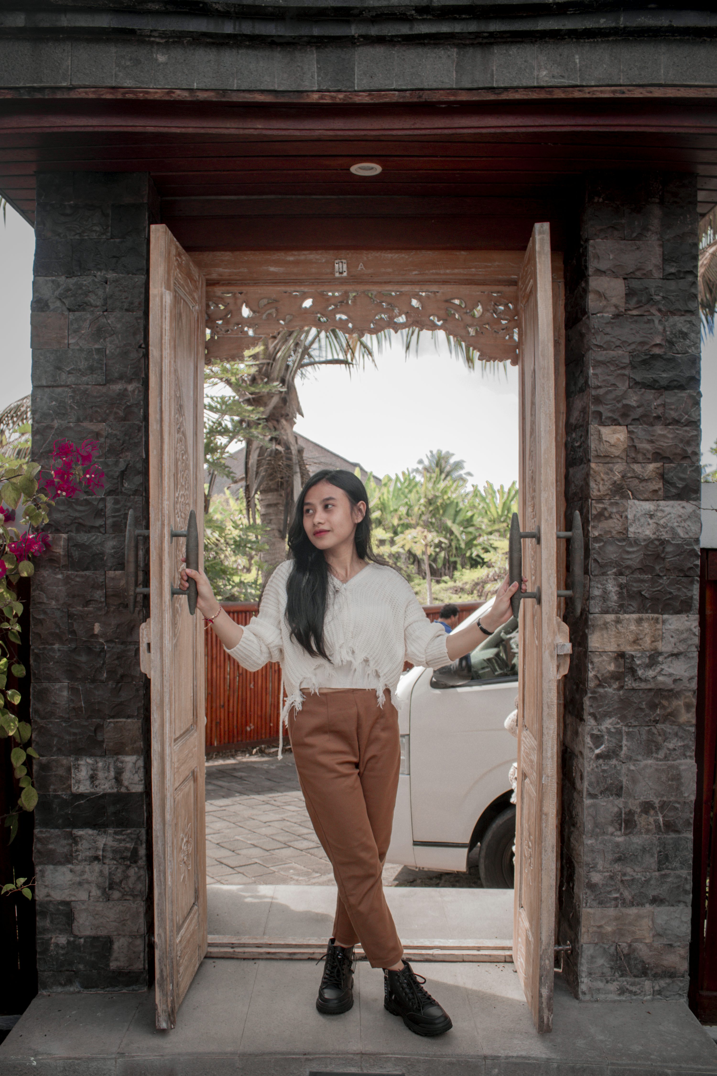 a young asian woman stands in a doorway, holding the handles of a pair of wooden doors. she's wearing a white sweater, brown pants, and black ankle boots. the background includes tropical plants, a white van, and a stone wall.  the scene is brightly lit, suggesting a daytime outdoor setting.  the architectural style hints at a possible balinese or indonesian location.  the overall feeling is relaxed and casual.