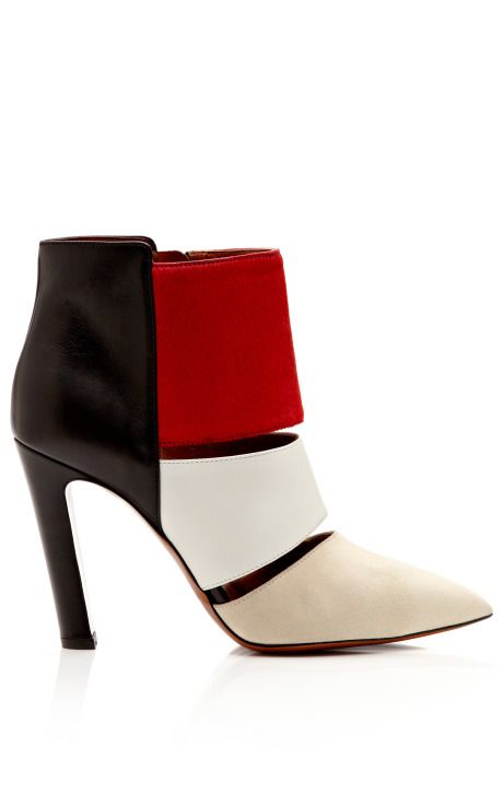 a stylish women's ankle boot with a high stiletto heel is displayed against a white background. the boot has a unique color block design, featuring black leather at the back of the heel and upper, a red suede panel above the ankle, a white leather strap around the midfoot, and a beige suede pointed toe. the boot has distinctive cutouts between the color blocks, showcasing an interesting and fashionable design. the high heel and pointy toe suggest a sophisticated and modern aesthetic.