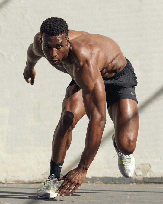a muscular young athlete in black athletic shorts and sneakers is running and posing for a shot, leaning down with his left leg bent, right leg stretched out, and his right hand reaching toward the camera. the athlete is shirtless, showing off his muscular physique. the shot is taken on a city street with a concrete wall in the background.  the athlete is looking determined and ready to run. this photo conveys the essence of athleticism, energy, and fitness.