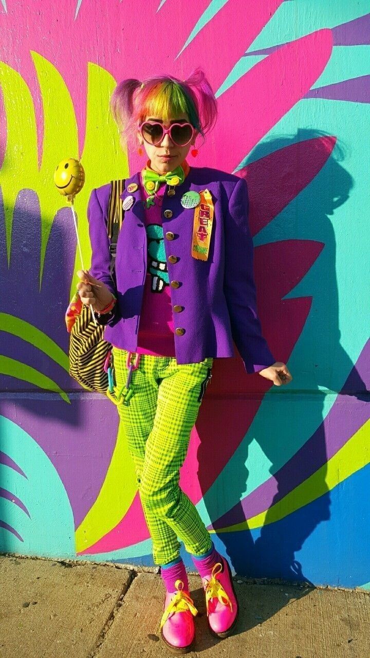a woman with rainbowcolored hair styled in pigtails stands in front of a vibrant, graffiticovered wall. she's wearing heartshaped sunglasses, a purple jacket adorned with badges, green plaid pants, and bright pink shoes with yellow laces. she holds a smiley face balloon and has a unique, eccentric style reminiscent of the 80s.