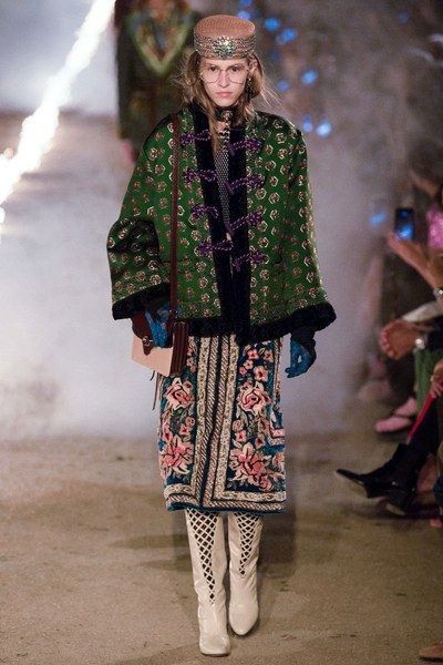 a full shot of a female model walking the runway at a gucci fashion show. she is wearing a green embroidered jacket, a floral embroidered skirt, white fishnet boots, a hat, and glasses. the background is blurred, showing other people and a runway setting. the overall atmosphere suggests a high fashion event.