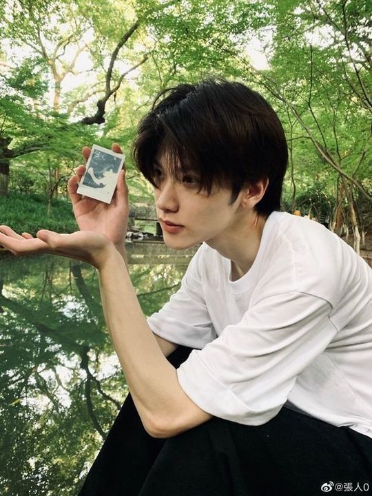 a young man with black hair and a white tshirt sits by a pond in a lush green forest. he holds a small photo in his left hand and gazes contemplatively at it. his reflection is visible in the calm water of the pond, which reflects the surrounding trees and foliage. the overall atmosphere is peaceful and serene, highlighting the beauty of the natural environment.
