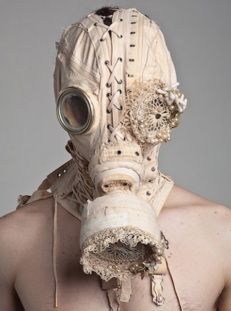 a person is wearing a white gas mask that is covered in lace and other decorative materials. the mask is over the person's head and is covering the face. the background is a plain light grey.