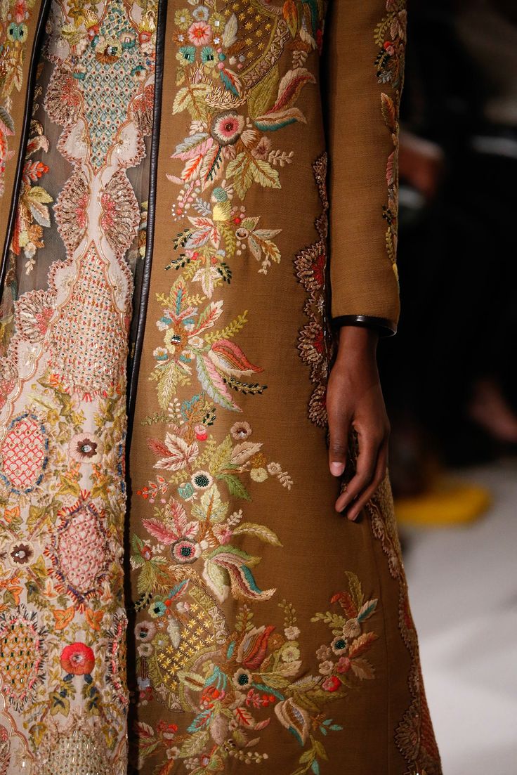 loseup view of a woman's long coat adorned with intricate floral embroidery. the embroidery is rich with detail, showcasing a variety of colorful flowers, leaves, and other botanical motifs. the coat is a muted brown color, and the embroidery uses a palette of autumnal shades.  the image focuses on the elaborate texture and craftsmanship of the embroidery. a portion of the under garment is also visible, suggesting a layered look. the model's hand is partially visible.