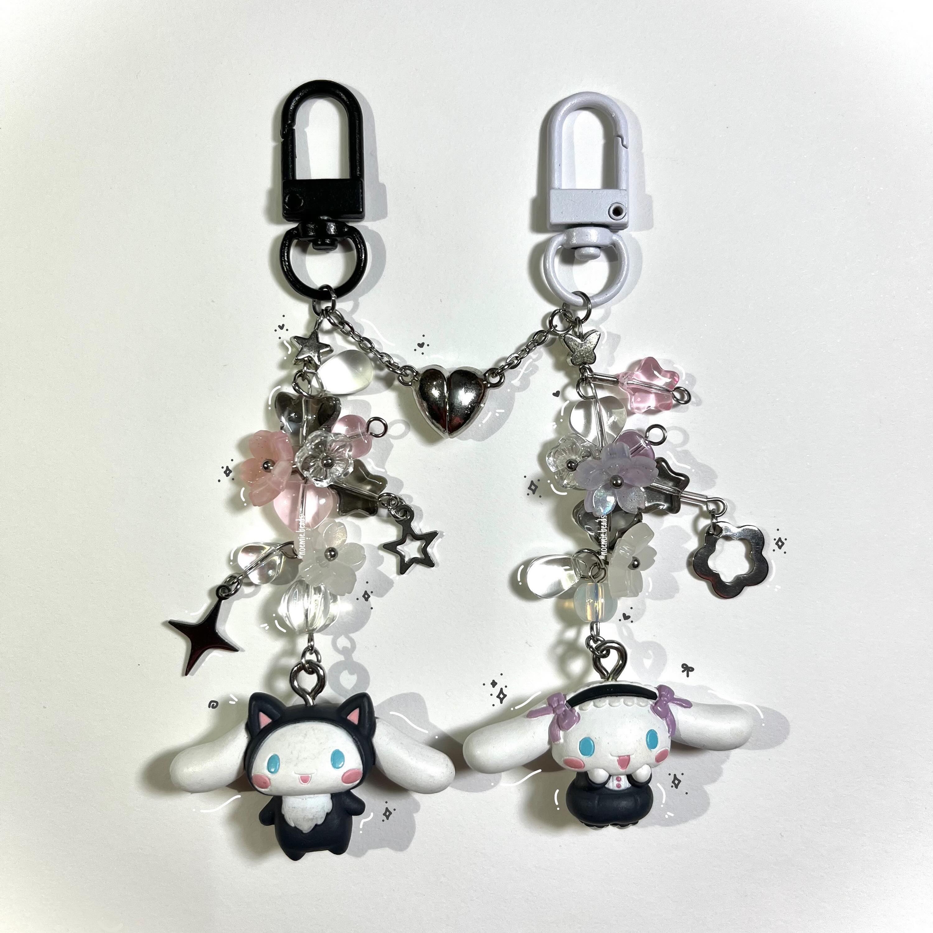 the image shows two cute, kawaii keychains. the left keychain features a black kuromi character, and the right one has a white cinnamoroll character. both are adorned with beads, flowers, and charms hanging from the clips. they are linked together by a chain with a heart in the middle.