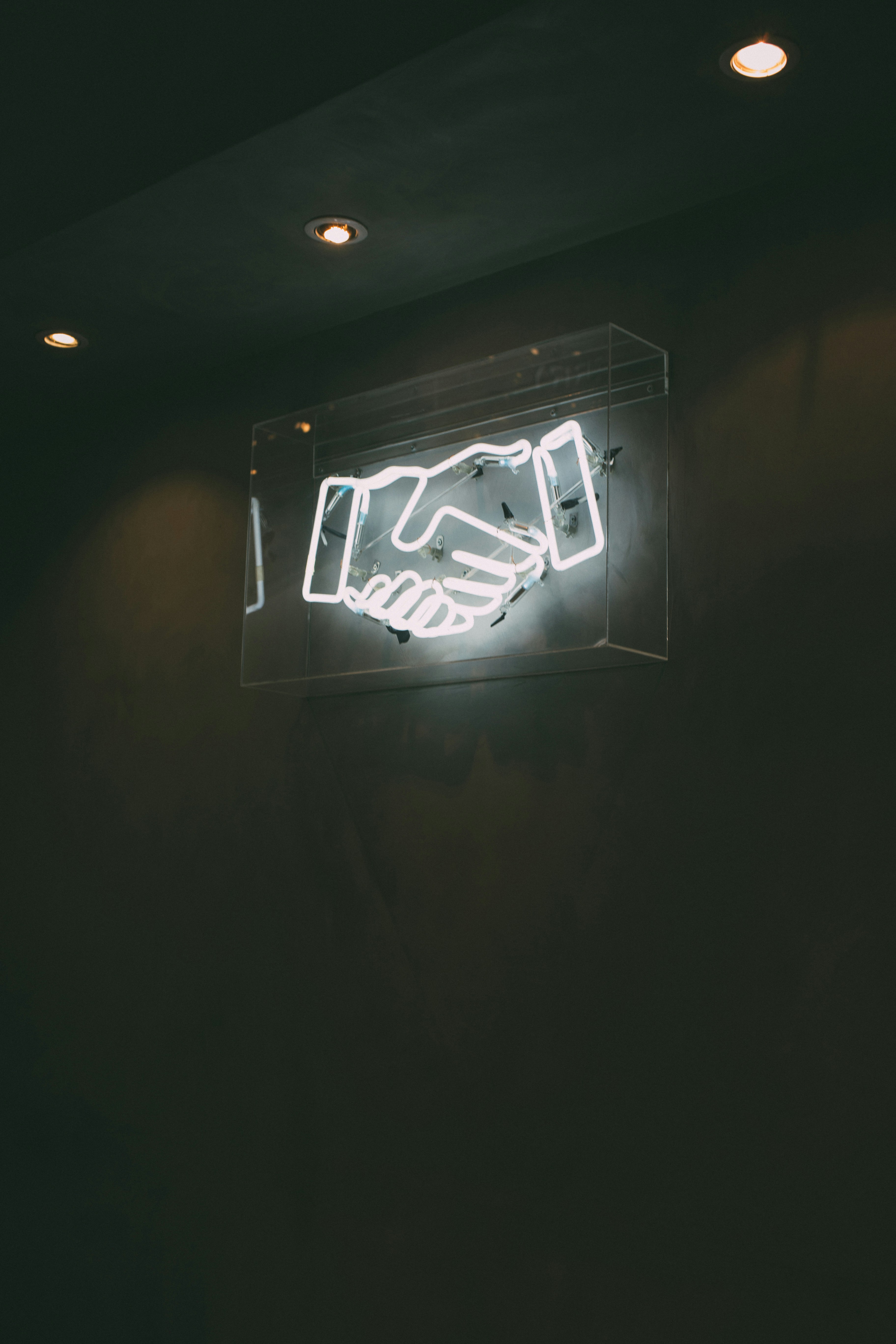 a neon sign of a handshake mounted on a dark wall. the sign is encased in a clear acrylic box and illuminated, creating a bright white glow against the dark background. three spotlights are positioned above the sign, casting subtle pools of light on the wall.