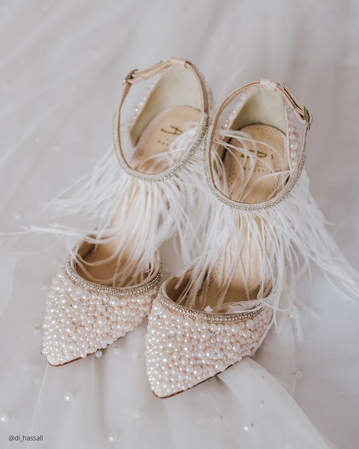 a pair of white bridal shoes with pearl embellishments and feather accents on a white background.