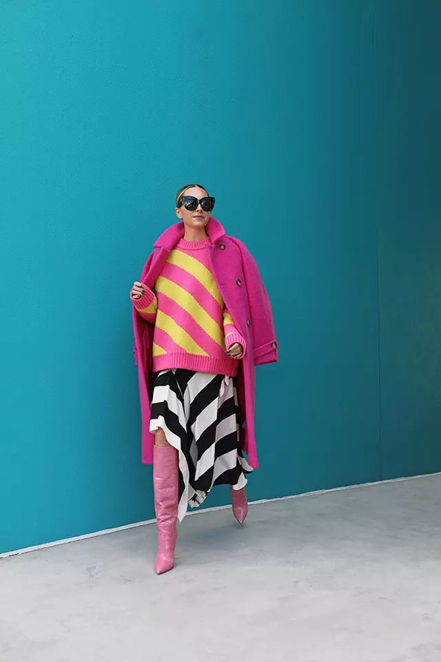 a fulllength shot of a woman standing in front of a teal wall. she is wearing a pink coat over a yellow and pink striped sweater, a black and white striped skirt, and pink boots. she is also wearing sunglasses.