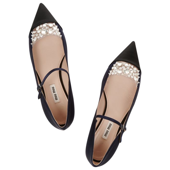 a pair of navy blue miu miu satin ballerinas with a pointed toe and black pointed toe cap.  the shoes feature a clear panel across the vamp with crystal embellishments. they have a delicate ankle strap with a buckle closure. the shoes are shown from an overhead perspective, and the miu miu branding is visible on the insole.