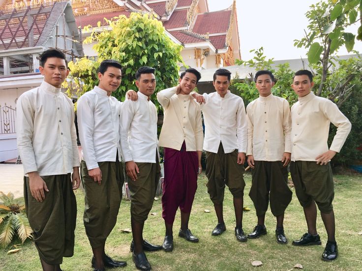 a group of seven young asian men are standing outdoors in front of a temple.  all men are smiling at the camera. they are wearing traditional white shirts with black pants and black footwear. the men are standing on a grassy area in front of the temple.