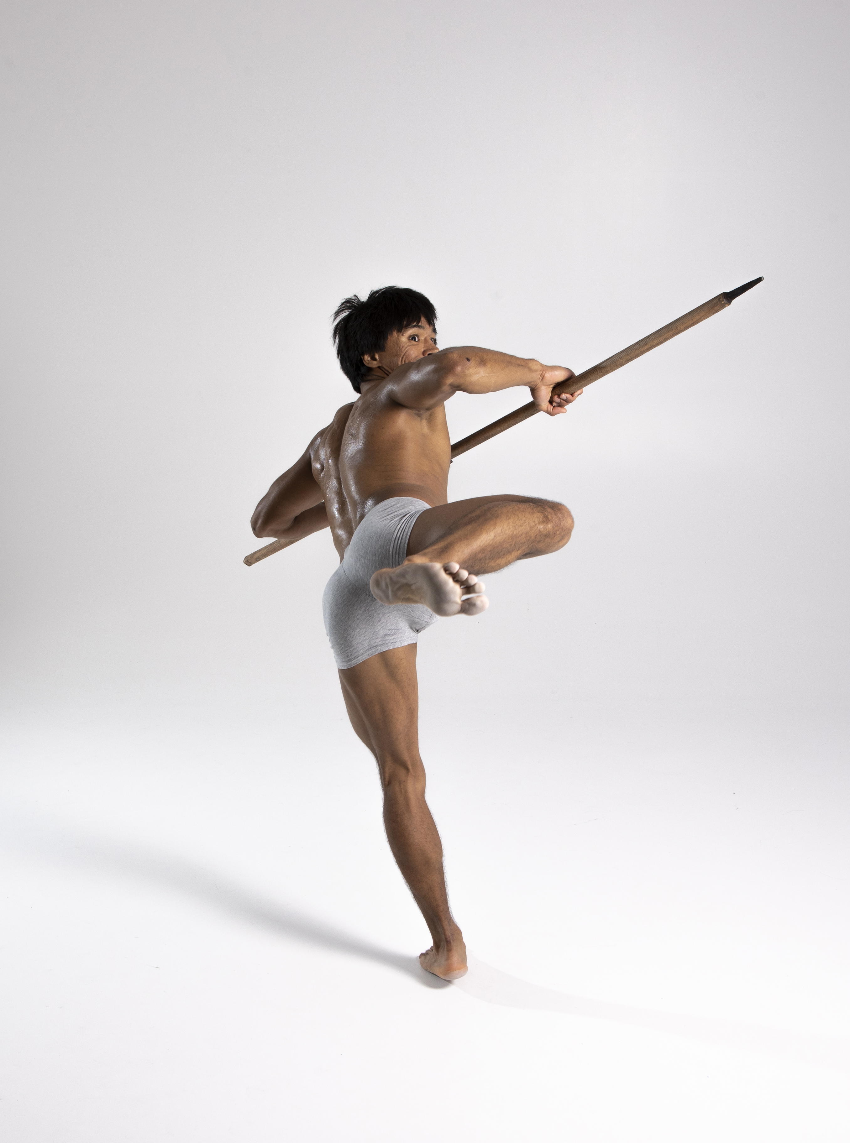 a muscular, shirtless man in gray shorts strikes a martial arts pose with a spear in a studio setting. he is barefoot, balanced on one leg, with the other knee raised. the spear is held horizontally, extending forward. the background is a neutral white. his body appears athletic and toned.