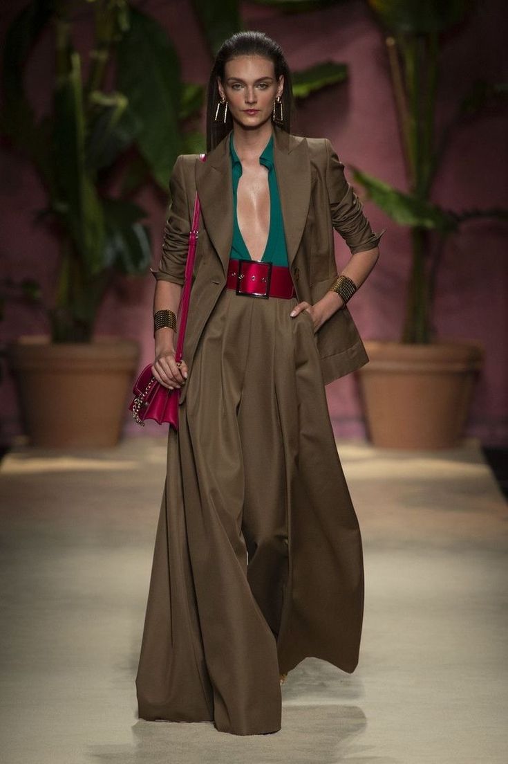 a full shot of a fashion model walking the runway. she is wearing a stylish brown pantsuit with wideleg trousers, an emerald green top underneath, a pink belt and matching handbag. she also sports long earrings and a bracelet. the background features lush green plants and a warmtoned wall. the overall ambiance is chic and elegant.