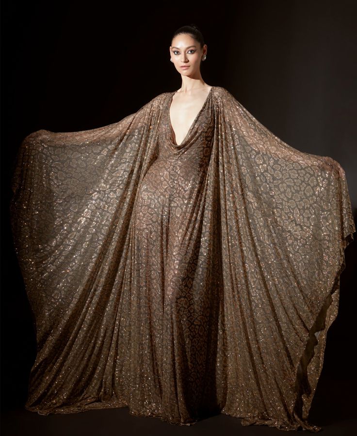 a female fashion model poses in a dramatic, floorlength gold sequin gown. the gown features a plunging neckline and a flowing, capelike silhouette. the model's hair is styled in a simple updo, and she wears minimal jewelry. the image is taken in a studio setting, with a dark background that highlights the gown's sparkle. the model appears confident and elegant in her attire.