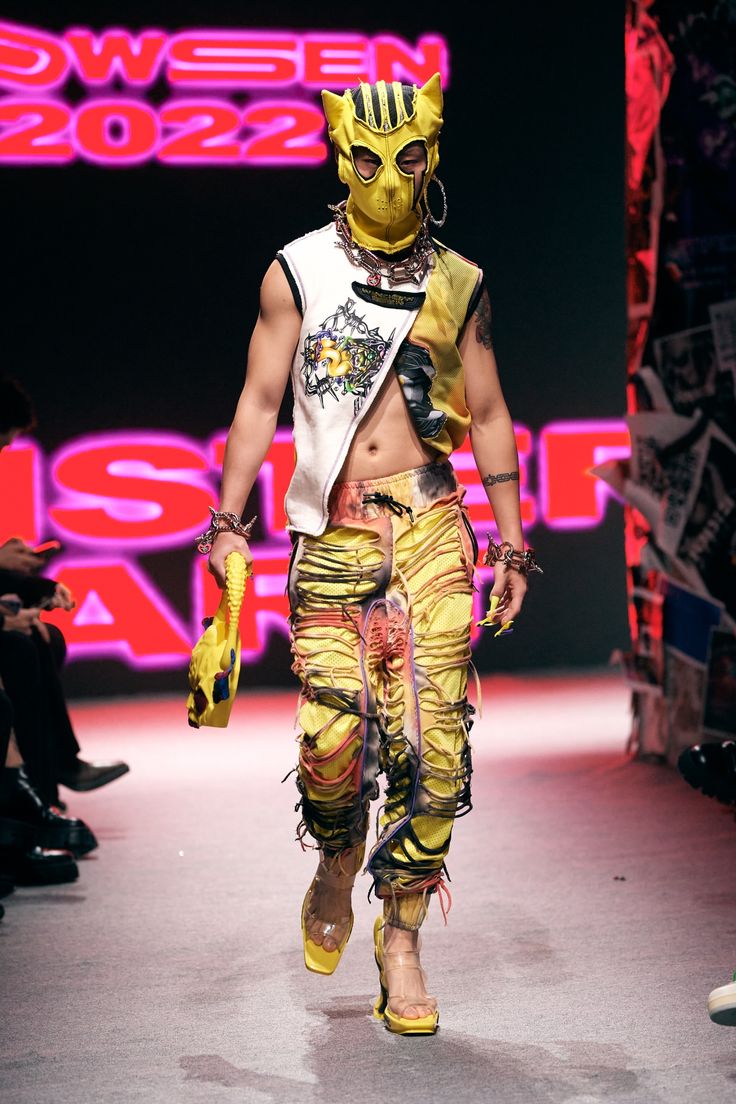 a fashion model walks the runway in a bold and stylish outfit. the model is wearing a yellow and black cat mask, a white sleeveless shirt, and yellow pants with intricate cutouts. the model is accessorizing with chains and rings and is sporting long nails, adding to their striking appearance. the background features a vibrant red neon sign and an array of graphic posters, enhancing the overall visual impact of the runway scene.
