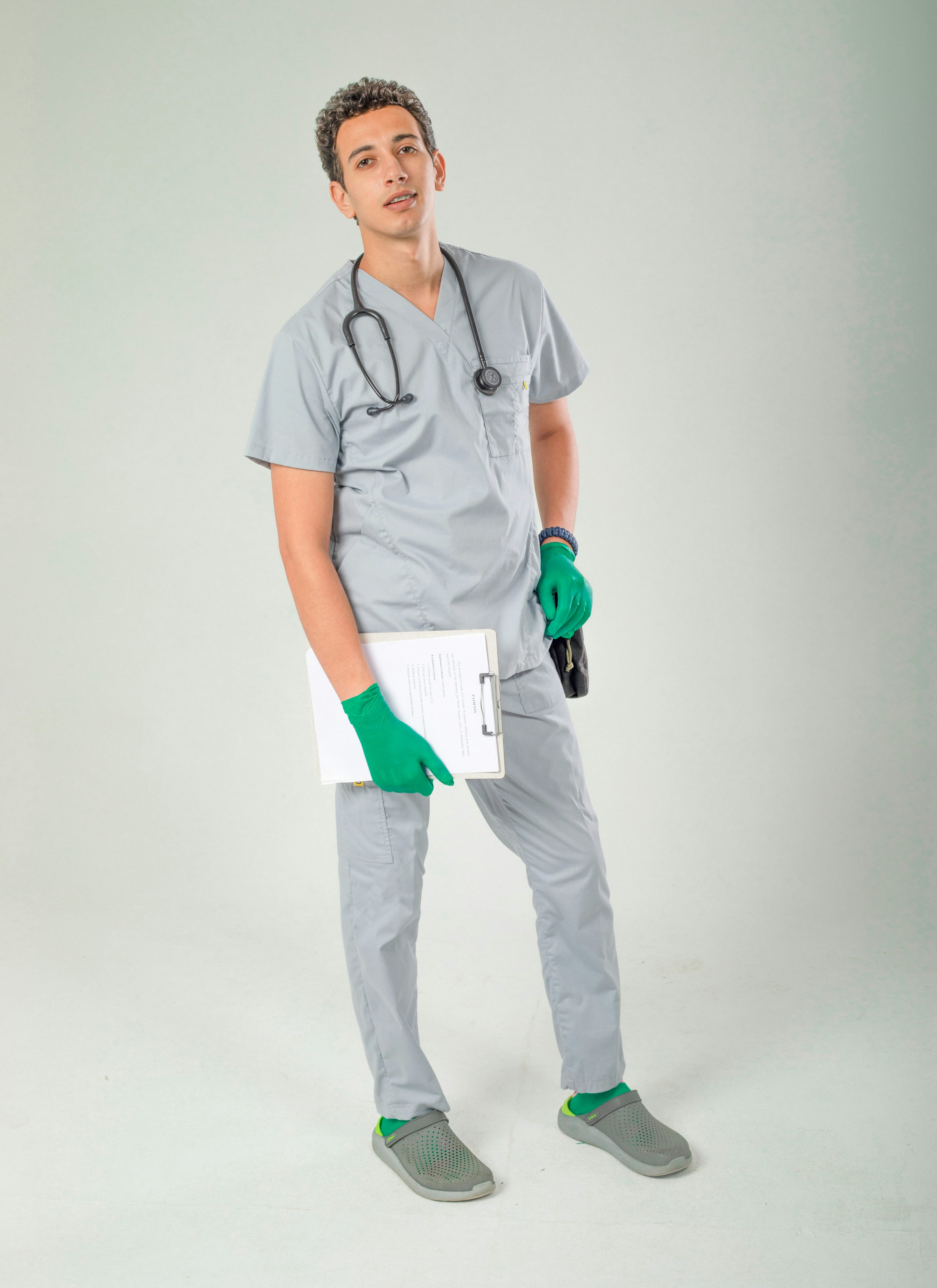 a fullbody studio shot of a young male doctor in grey scrubs, wearing a stethoscope around his neck, holding a clipboard with medical records and wearing green gloves. he's standing confidently against a light grey background, and his shoes are grey clogs.  the overall impression is one of professionalism and readiness.