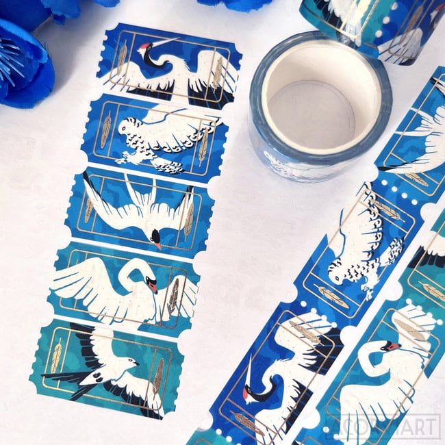 a closeup shot shows two rolls of washi tape featuring a repeating pattern of various birds on a teal background. one roll is partially unrolled, showcasing the design, while the other roll is still fully wound. the birds depicted include cranes, swans, and owls, all rendered in white and gold against the blue backdrop. the tape has a ticketlike border, adding a vintage aesthetic. the image is welllit and the details of the tape are sharp. blue flowers are visible in the upper left corner, providing a contrasting color element. the overall impression is one of delicate beauty and artistic design.