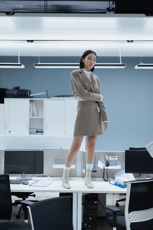 a young asian woman in a beige suit and white socks and boots stands confidently on a modern office desk. the office is minimalist with white and grey tones.  computers and office equipment are visible, but the focus remains on the woman. the overall style is clean and professional.