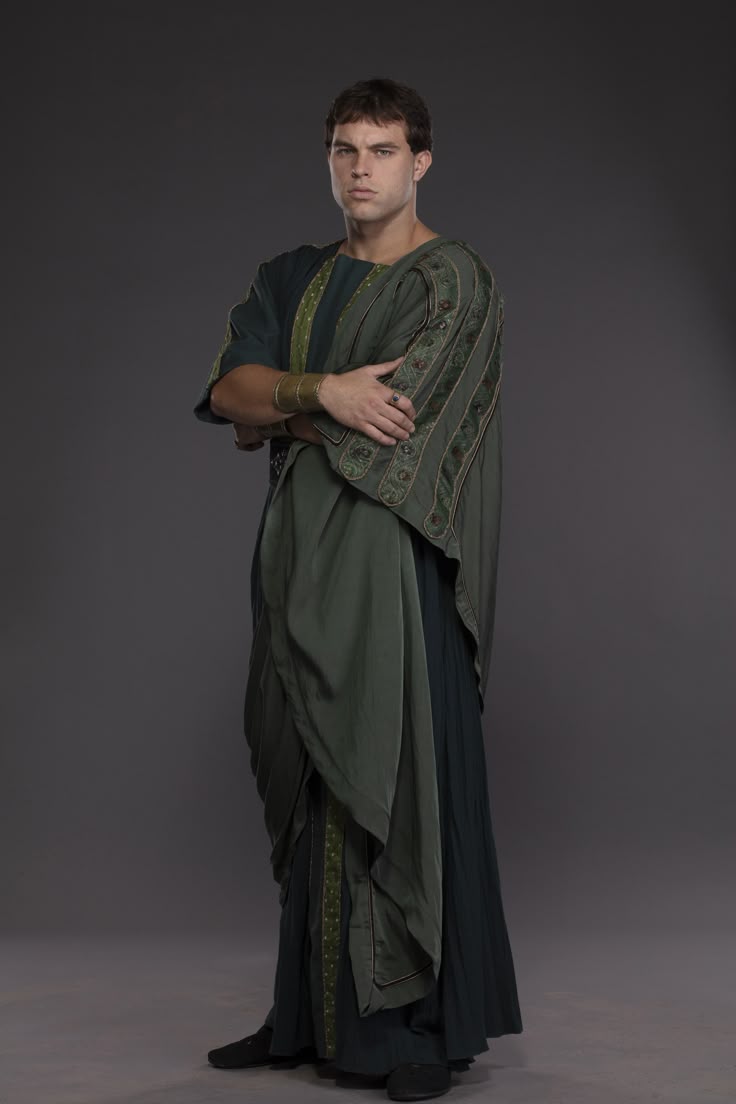 a full shot shows a seriouslooking man in a roman toga, standing in a studio against a dark background with his arms crossed. he is wearing a green and dark green toga over a tunic, accessorized with a golden arm cuff and a ring on his left hand. he is positioned in the center of the image, looking directly at the camera.
