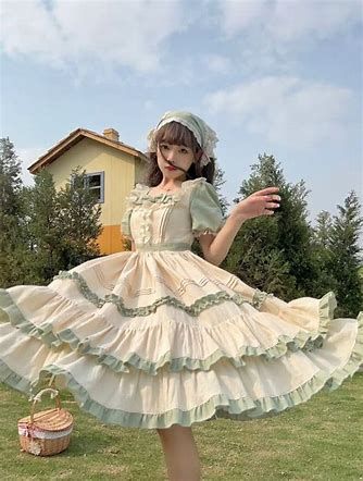 a young woman in a vintage style dress is posing in a field. she is wearing a green and cream colored dress with lace and ruffles. the dress is long and flowy, and the skirt is tiered. the woman has long brown hair and is wearing a white headscarf. the background of the image is a green field with a house in the distance. the lighting is bright and sunny, and the overall feeling of the image is happy and carefree.