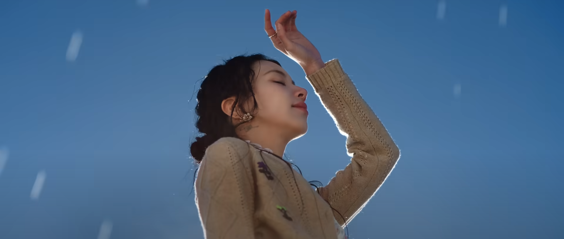 a young woman, with her eyes closed, stands with her face towards the blue sky and one arm raised.  snowflakes are gently falling around her. the scene is bathed in soft sunlight.