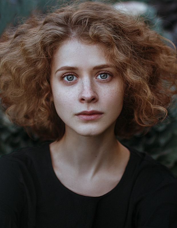 a portrait of a woman with red curly hair and freckles. she has light blue eyes and a neutral expression. she is wearing dark clothing and has fair skin. the background is blurred.