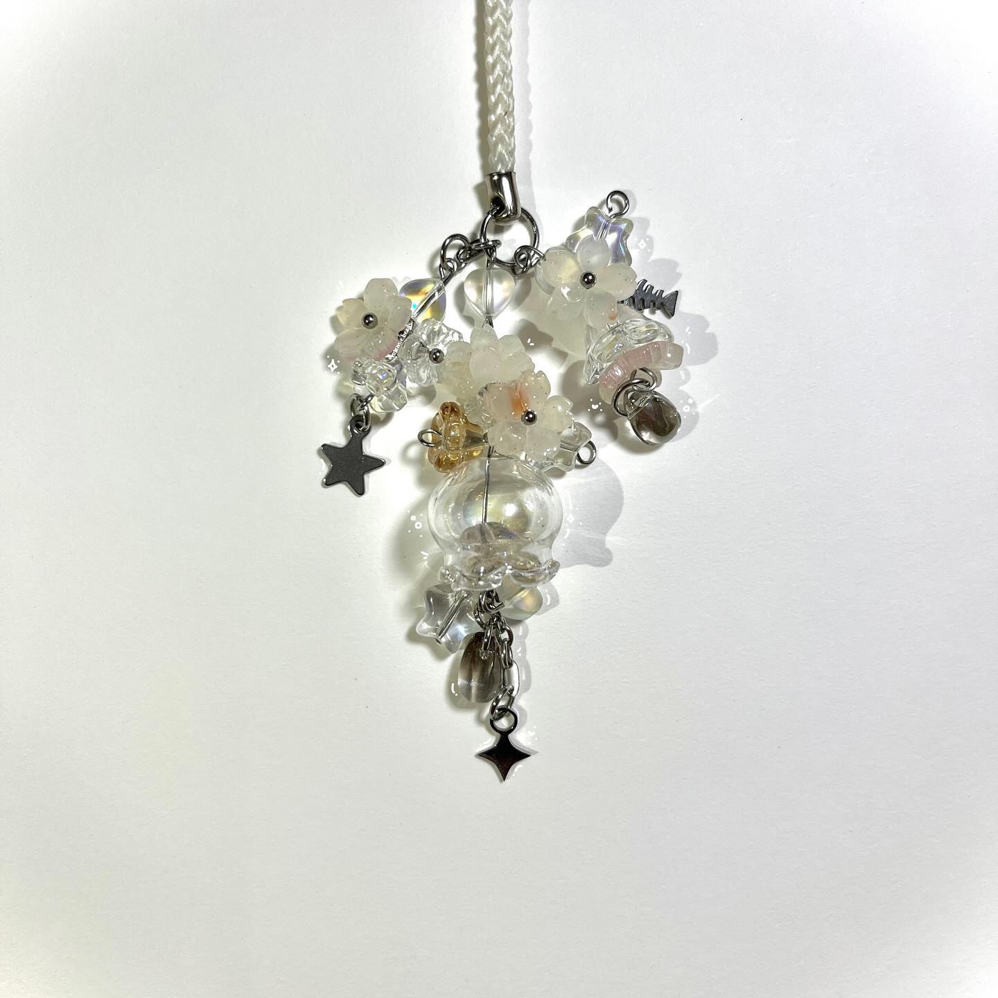a closeup shot showcases a handmade charm, possibly for a mobile phone or as a pendant. the charm features a cluster of translucent white flower beads, interspersed with clear and iridescent glass beads. a small, spherical glass container with a few beads inside is centrally located. metallic charms in the shape of stars and other abstract forms dangle from the lower portion of the charm, connected by delicate chains. the charm is suspended from a white braided cord attached to a silver clasp, against a plain white backdrop.