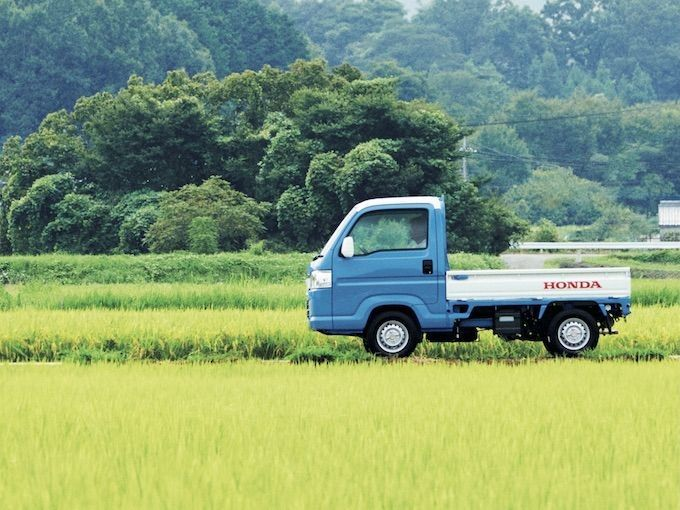 the image shows a blue and white honda acty kei truck parked in a green rice field in a rural area, possibly in japan. the truck is a compact vehicle designed for light transportation. in the background, there are trees and a hazy sky.
