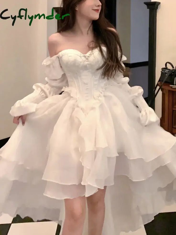 a woman poses indoors wearing a white offtheshoulder dress with a corset bodice, long ruffled sleeves, and a highlow hem with multiple layers of ruffles.