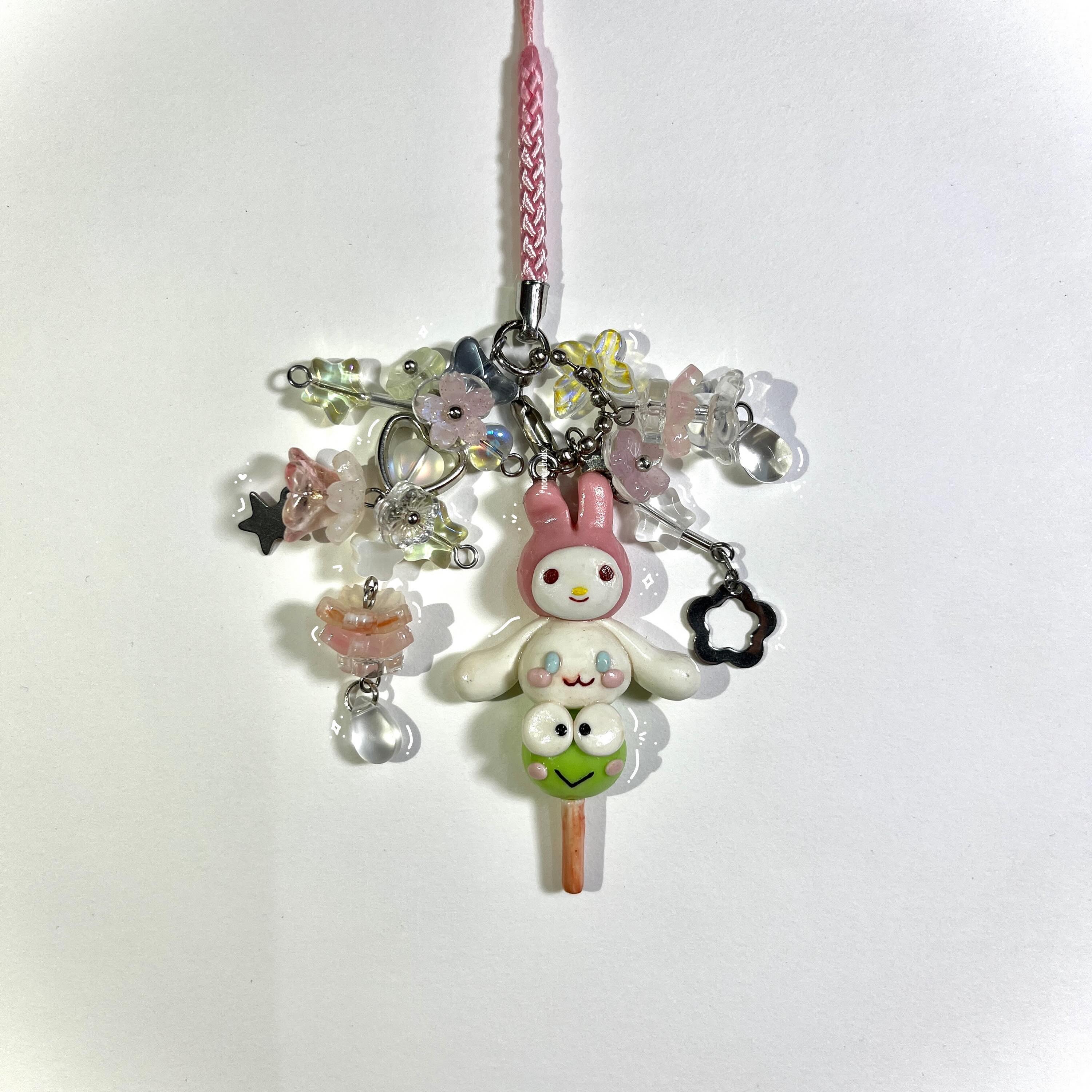 a closeup of a handmade keychain featuring a my melody character stacked on top of a keroppi character, surrounded by various colorful beads, flower and star charms. the keychain is attached to a pink braided cord and has a silver clasp.