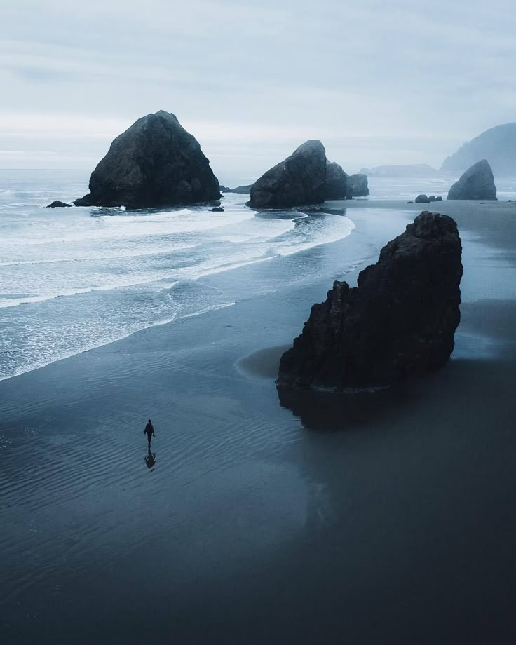the image shows a person walking on a dark, wet sandy beach with several large rock formations in the background and along the shoreline. the water is reflecting the sky, creating a moody and somewhat lonely atmosphere. the sky is overcast and grey, adding to the somber tone.