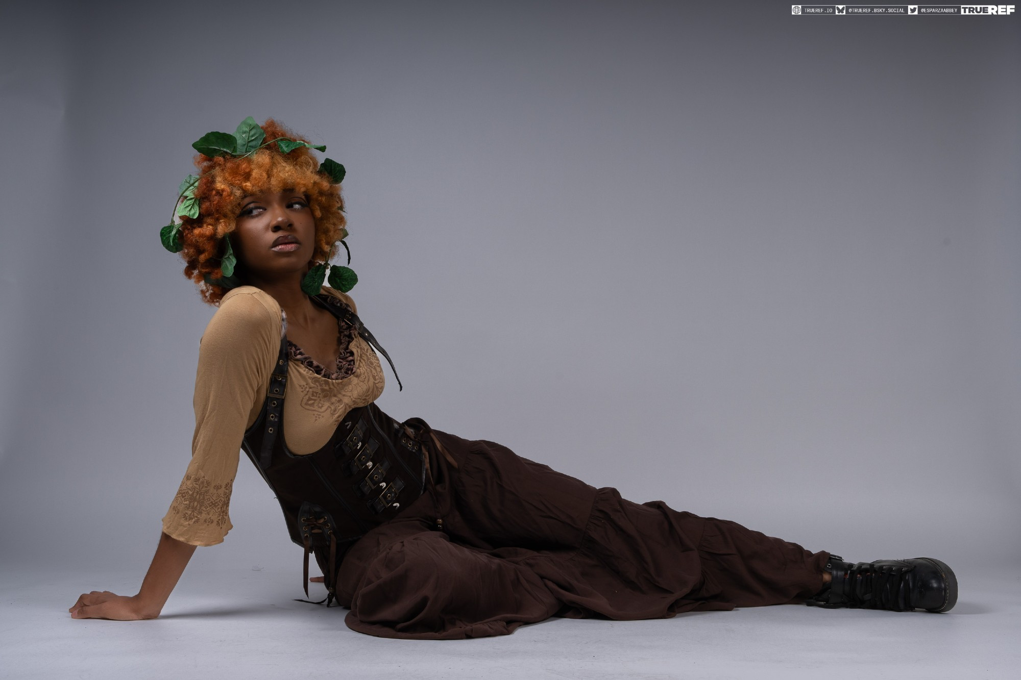 a young woman with curly orange hair adorned with green leaves lies on a grey studio floor.  she is wearing a brown, steampunkinspired outfit consisting of a corset and loosefitting pants, and black boots. her pose is relaxed, and she looks away from the camera. the overall style is a blend of fantasy and fashion.